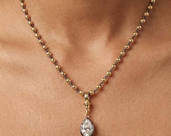 May include: A gold-toned necklace with a teardrop-shaped pendant. The necklace features small, round beads and a pendant with a clear, sparkling stone. The pendant is set in a dark metal frame, creating a contrast with the gold chain.