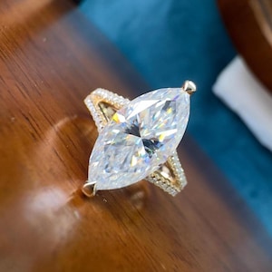 Puede incluir: Un anillo de compromiso con diamante talla marquesa y banda dorada. El diamante grande y transparente es el punto focal, engastado en una montura de garras. La banda presenta piedras de acento más pequeñas, agregando brillo. El anillo se exhibe sobre una superficie de madera.