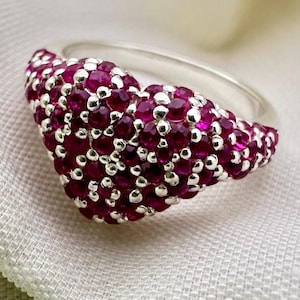 May include: A silver ring featuring a heart-shaped setting densely encrusted with numerous small, round, deep pinkish-red gemstones. The stones are set closely together in a pavé style, covering the entire top surface of the heart-shaped metal band.