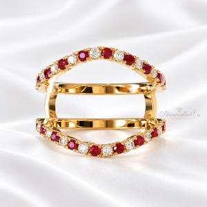 Könnte beinhalten: Ein goldener Doppelring mit zwei geschwungenen Reihen aus abwechselnd runden roten Rubinen und klaren weißen Diamanten. Die beiden Ringbänder sind an den Seiten durch vertikale Goldstege verbunden, was ein offenes, geschichtetes Design ergibt.