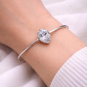 May include: A silver-toned bracelet featuring a pear-shaped, clear gemstone. The gemstone is the focal point, set on a delicate, thin band. The bracelet is shown on a wrist, against a soft, white background.