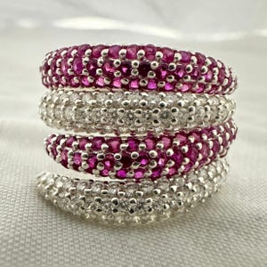 Könnte beinhalten: Ein mehrreihiger Bypass-Ring mit abwechselnden Bändern aus pavé-gefassten runden rosa Rubinen und klaren weißen Diamanten. Das Design besteht aus zwei Reihen leuchtend rosa Steinen und zwei Reihen funkelnder weißer Steine in poliertem Metall.