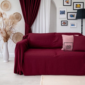 Peut inclure: Une housse de canapé en lin bordeaux avec deux coussins, un rose et un autre avec le texte "LINEN SHADES". Le canapé est dans un salon avec des murs blancs et un rideau blanc derrière.