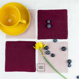 Op de afbeelding: Vier vierkante linnen onderzetters in bordeauxrood en blush roze met een gele narcissen en blauwe bessen erop. De onderzetters zijn gemaakt door Linen Shades.