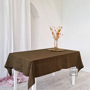 May include: A brown linen tablecloth on a white table with a vase of dried flowers and a woven placemat. The tablecloth is made of natural linen and has a rustic look. The text "LINEN SHADES" is visible in the lower right corner of the image.