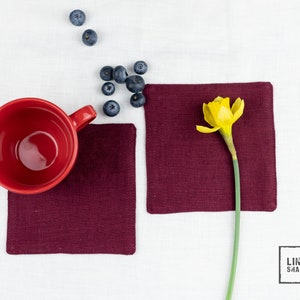 Könnte beinhalten: Zwei burgunderfarbene Untersetzer aus Leinen mit einer roten Keramiktasse und einer gelben Narzisse auf einem weißen Leinenhintergrund. Der Text "LINEN SHADES" ist in Schwarz auf dem weißen Leinen gedruckt.