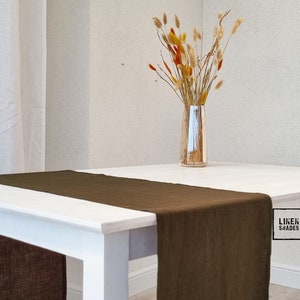 Può includere: Un runner da tavolo in lino marrone su un tavolo bianco con un vaso di fiori secchi. Il runner da tavolo è realizzato in lino naturale e ha una texture delicata. Il vaso è in vetro trasparente e i fiori sono un mix di beige, marrone e arancione. Il testo "LINEN SHADES" è visibile sul tavolo.