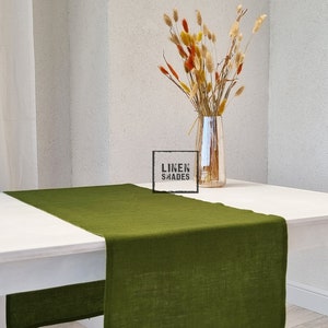 May include: A white table with a green linen table runner. The table runner is made of natural linen and has a subtle texture. The table is set with a vase of dried flowers.