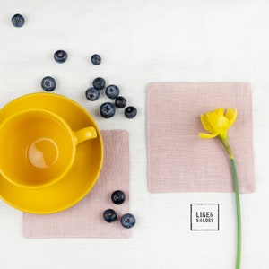 Könnte beinhalten: Eine gelbe Teetasse und Untertasse aus Keramik auf einem rosa Leinen-Platzset mit einer einzelnen gelben Narzisse auf einem rosa Leinen-Platzset. Blaubeeren sind um die Platzsets herum verstreut.