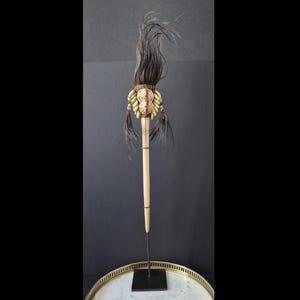 Könnte beinhalten: Ein dekorativer Stab mit einem langen, hellen Holzschaft, der mit einer Ansammlung cremefarbener Muscheln, dunkelbraunen Federn und einem hohen, aufrechten Büschel aus dunklen, haarähnlichen Fasern verziert und auf einem schwarzen Metallständer montiert ist.