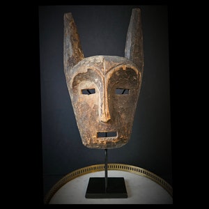 May include: A wooden mask with a carved face and large ears, displayed on a black stand. The mask is brown and has a weathered appearance.