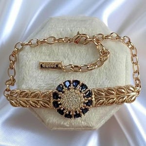Könnte beinhalten: Ein goldfarbenes Armband mit einem zentralen kreisförmigen Blumenmotiv, das aus einer Ansammlung klarer Kristalle besteht, umgeben von dunkelblauen tropfenförmigen Steinen. Das Band besteht aus zwei symmetrischen Blattmustern und einer Kette mit einem GUESS-Anhänger.