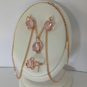 Könnte beinhalten: Ein Schmuckset aus Roségold, bestehend aus einer Halskette, einem Paar Ohrringen und einem Ring. Jedes Stück verfügt über einen großen, oval geschliffenen, blassrosa Edelstein, der von kleinen, klaren, funkelnden Steinen umgeben ist.
