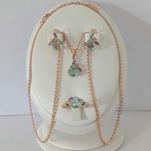 Könnte beinhalten: Ein Schmuckset aus Roségold bestehend aus einer Halskette, einem Paar Ohrringen und einem Ring. Jedes Stück verfügt über einen zentralen hellgrünen birnenförmigen Stein, akzentuiert durch kleine blaue und violette Steine im Marquise-Schliff.