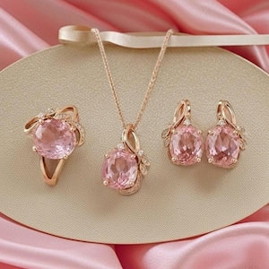 Può includere: Un set di gioielli composto da un anello, una collana con pendente e un paio di orecchini in oro rosa. Ogni pezzo presenta una grande gemma rosa chiaro dal taglio ovale, impreziosita da piccole pietre trasparenti incastonate nel metallo.