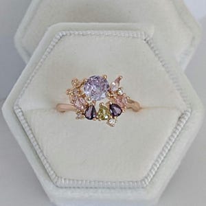 Könnte beinhalten: Ein roségoldener Ring mit einem großen, runden, hellvioletten Edelstein, umgeben von kleineren farbigen Edelsteinen in Rosa, Lila und Grün. Der Ring hat ein Cluster-Design mit kleinen Akzentsteinen.