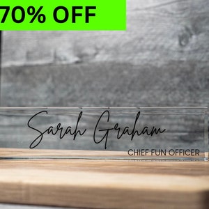 May include: A clear acrylic desk nameplate with the name "Sarah Graham" and the title "CHIEF FUN OFFICER" in black script. A bright green rectangle with the text "70% OFF" is in the upper left corner. The nameplate sits on a wooden surface.