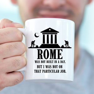 May include: White ceramic mug with a black graphic of a building with columns and two figures digging. The text "ROME WAS NOT BUILT IN A DAY. BUT I WAS NOT ON THAT PARTICULAR JOB." is printed below the graphic.