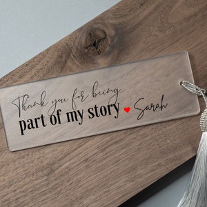 May include: Clear acrylic bookmark with a white tassel. The bookmark has the text "Thank you for being part of my story" with a red heart and the name "Sarah" written in cursive.