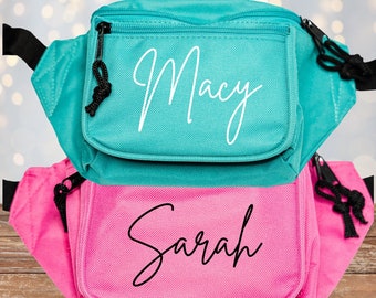 May include: Two personalized fanny packs, one teal and one pink. The teal pack has the name "Macy" in white script, and the pink pack has "Sarah" in white script. Both have black zippers and straps, perfect for carrying essentials.
