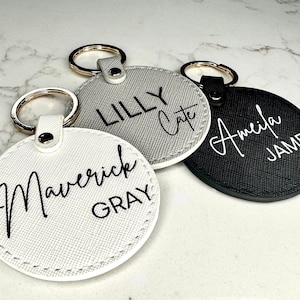 May include: Three round leather keychains with gold key rings. The keychains are white, gray, and black. The white keychain has the text "Maverick Gray" in black script. The gray keychain has the text "Lilly Cate" in black script. The black keychain has the text "Amelia James" in white script.
