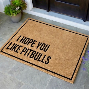 May include: A rectangular doormat made of natural coir fiber. The mat features the text "I HOPE YOU LIKE PITBULLS" in bold black letters. A black border frames the text and the mat itself. The doormat is placed on a gray tiled surface.
