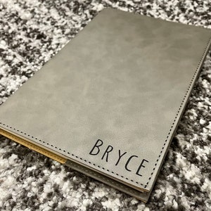 May include: A gray faux leather journal with a stitched edge and the name "Bryce" embossed in black lettering.
