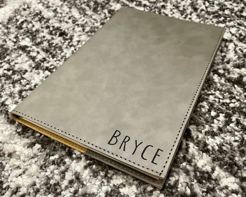 May include: A gray faux leather journal with a stitched edge and the name "Bryce" embossed in black lettering.