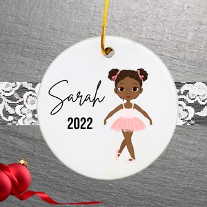 May include: A white ceramic Christmas ornament featuring a cartoon illustration of a young Black girl wearing a pink tutu and ballet shoes. The ornament is personalized with the name "Sarah" and the year "2022".