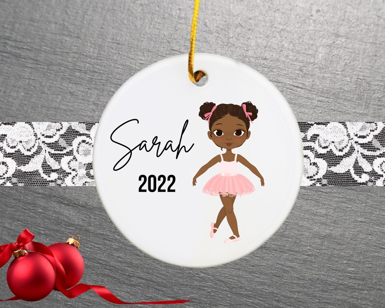 May include: A white ceramic Christmas ornament featuring a cartoon illustration of a young Black girl wearing a pink tutu and ballet shoes. The ornament is personalized with the name "Sarah" and the year "2022".
