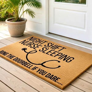May include: A rectangular doormat with the text "NIGHT SHIFT NURSE SLEEPING RING THE DOORBELL IF YOU DARE..." in black. A black stethoscope graphic is also featured. The mat is tan and sits on a wooden porch.