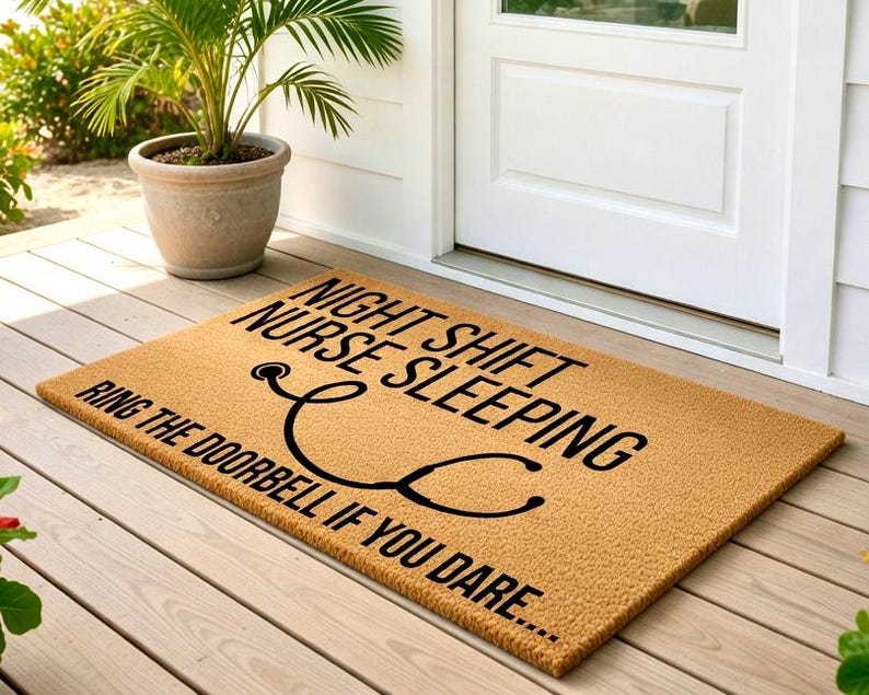 May include: A rectangular doormat with the text "NIGHT SHIFT NURSE SLEEPING RING THE DOORBELL IF YOU DARE..." in black. A black stethoscope graphic is also featured. The mat is tan and sits on a wooden porch.