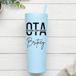 Puede incluir: Un vaso de color azul claro con el texto "OTA OCCUPATIONAL THERAPY ASSISTANT" y el nombre "Britney" en negro. Tiene una tapa a juego y una pajita azul. El fondo es una superficie de madera blanca.