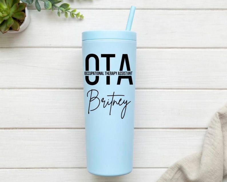 Puede incluir: Un vaso de color azul claro con el texto "OTA OCCUPATIONAL THERAPY ASSISTANT" y el nombre "Britney" en negro. Tiene una tapa a juego y una pajita azul. El fondo es una superficie de madera blanca.