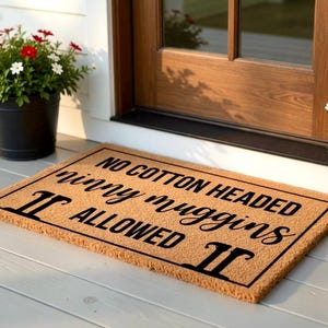 Pode incluir: Um tapete retangular com o texto "NO COTTON HEADED ninny muggins ALLOWED" em letras pretas. O tapete &eacute; de cor castanho claro e est&aacute; colocado em frente a uma porta de madeira. &Agrave; esquerda, h&aacute; um vaso preto com flores vermelhas e brancas.