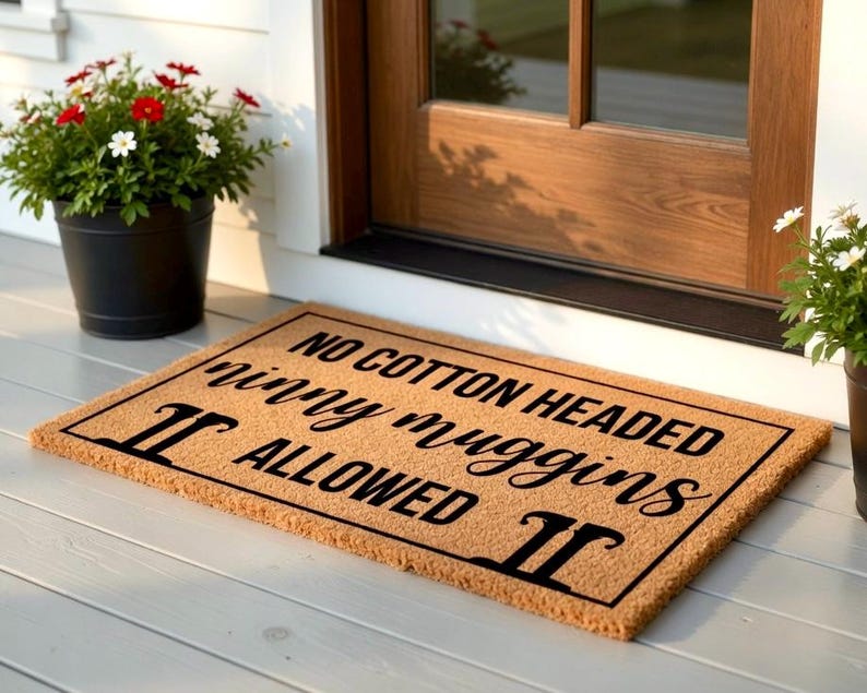 Pode incluir: Um tapete retangular com o texto "NO COTTON HEADED ninny muggins ALLOWED" em letras pretas. O tapete &eacute; de cor castanho claro e est&aacute; colocado em frente a uma porta de madeira. &Agrave; esquerda, h&aacute; um vaso preto com flores vermelhas e brancas.
