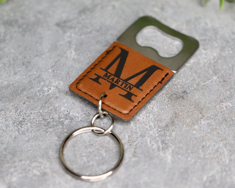 Op de afbeelding: Een bruine leren sleutelhanger met een flesopener en een metalen ring. Het leer is gegraveerd met de letter "M" en de naam "Martin".