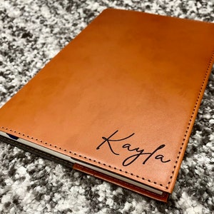 May include: A brown leather journal with a stitched edge and the name "Kayla" engraved on the front cover.