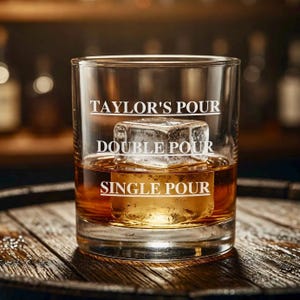 May include: A rocks glass with the text "TAYLOR'S POUR", "DOUBLE POUR", and "SINGLE POUR" etched on the glass. The glass contains a large ice cube and amber-colored liquid, resting on a wooden surface.
