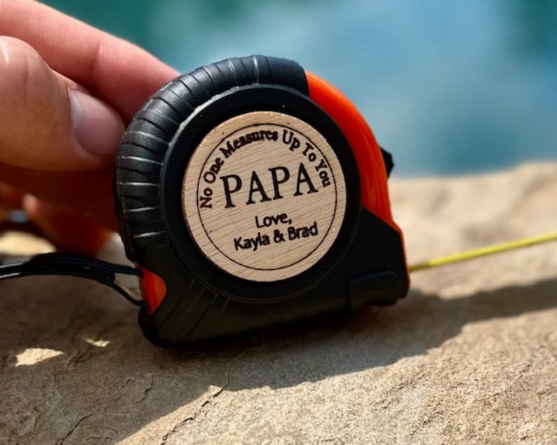 May include: A black and orange tape measure with a wooden circle that says "No One Measures Up To You Papa Love, Kayla & Brad"