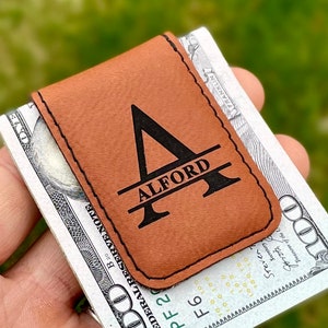 May include: A brown leather money clip with a black engraved initial "A" and the name "ALFORD" underneath. The money clip is holding a US dollar bill.