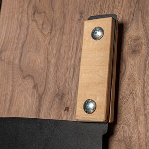 Puede incluir: Primer plano de un mango de madera con dos tornillos plateados unidos a un marco de metal negro. El mango es de madera marr&oacute;n claro y tiene una superficie lisa.