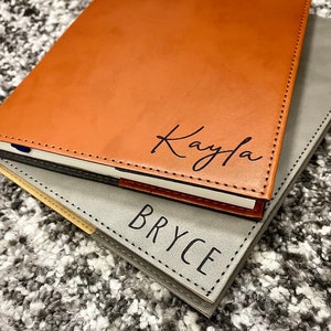 May include: Two leather-bound journals, one brown with the name "Kayla" embossed in black, and the other gray with the name "Bryce" embossed in black.