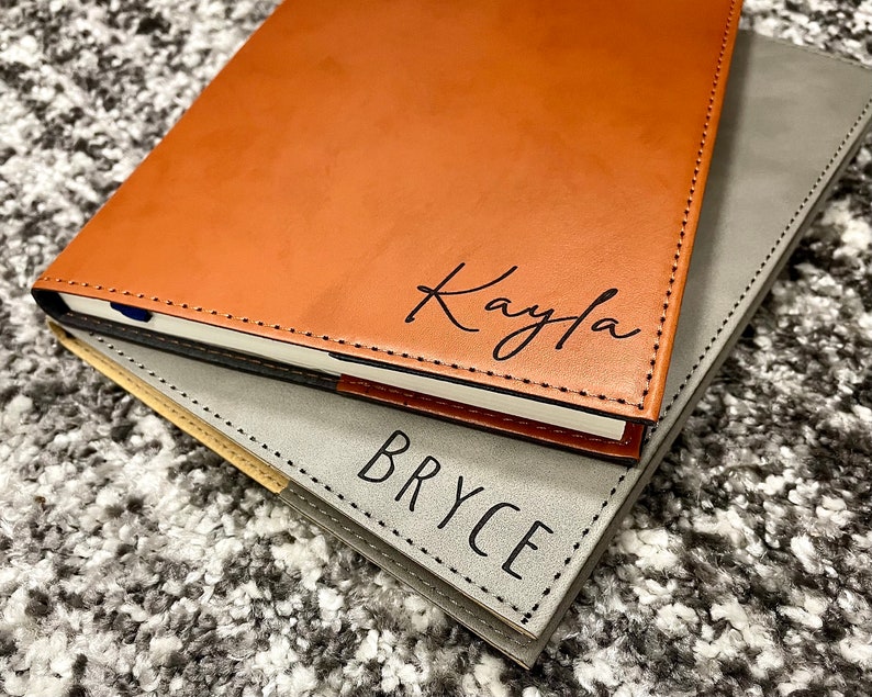 May include: Two leather-bound journals, one brown with the name "Kayla" embossed in black, and the other gray with the name "Bryce" embossed in black.