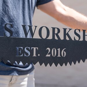 Puede incluir: Un letrero de metal negro con el texto "Woods Workshop Est. 2016" en forma de sierra.