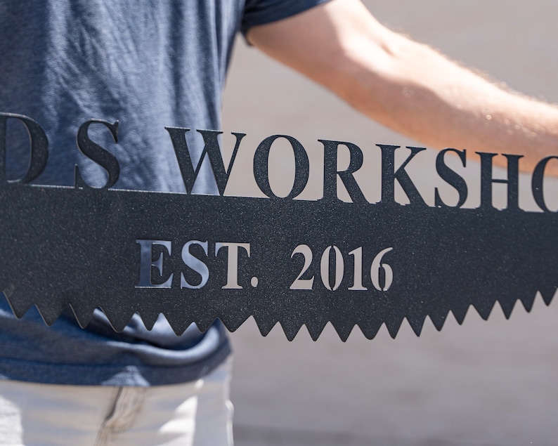 Puede incluir: Un letrero de metal negro con el texto "Woods Workshop Est. 2016" en forma de sierra.