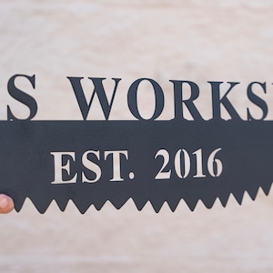 Puede incluir: Un letrero de metal negro con las palabras "Wood's Workshop Est. 2016" en letras blancas. El letrero tiene forma de hoja de sierra.
