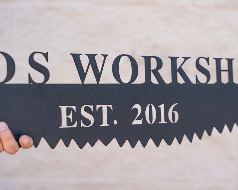 Puede incluir: Un letrero de metal negro con las palabras "Wood's Workshop Est. 2016" en letras blancas. El letrero tiene forma de hoja de sierra.