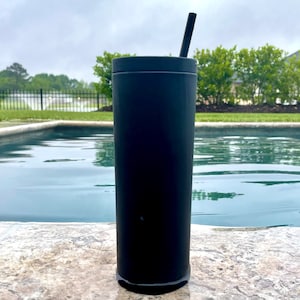 May include: A black stainless steel tumbler with a straw, sitting on a stone surface by a swimming pool.
