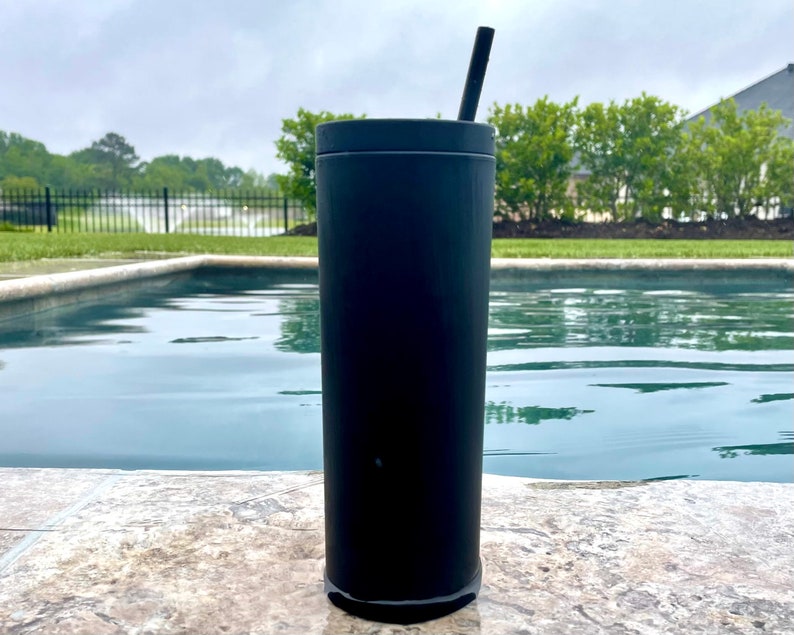 May include: A black stainless steel tumbler with a straw, sitting on a stone surface by a swimming pool.